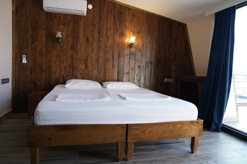 Una cama bien hecha con sábanas blancas en una habitación con paneles de madera y un amplio ventanal con cortinas azules.