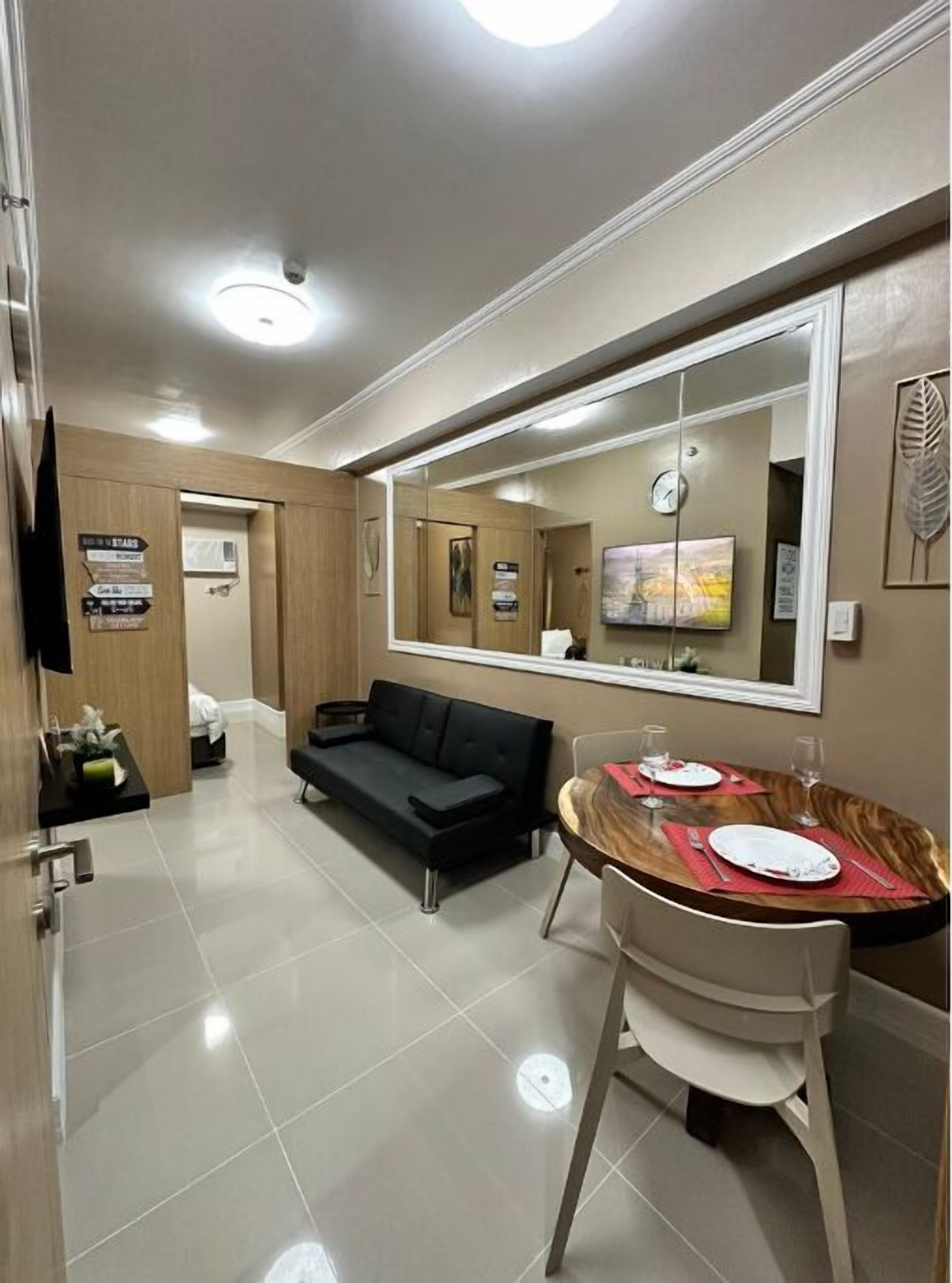 A modern hotel room with a black sofa, a dining table with red placemats, and a large mirror reflecting the room.
