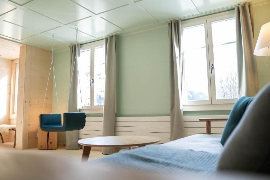 Una habitación con cama, una silla colgante, una mesa redonda y ventanas grandes con cortinas.