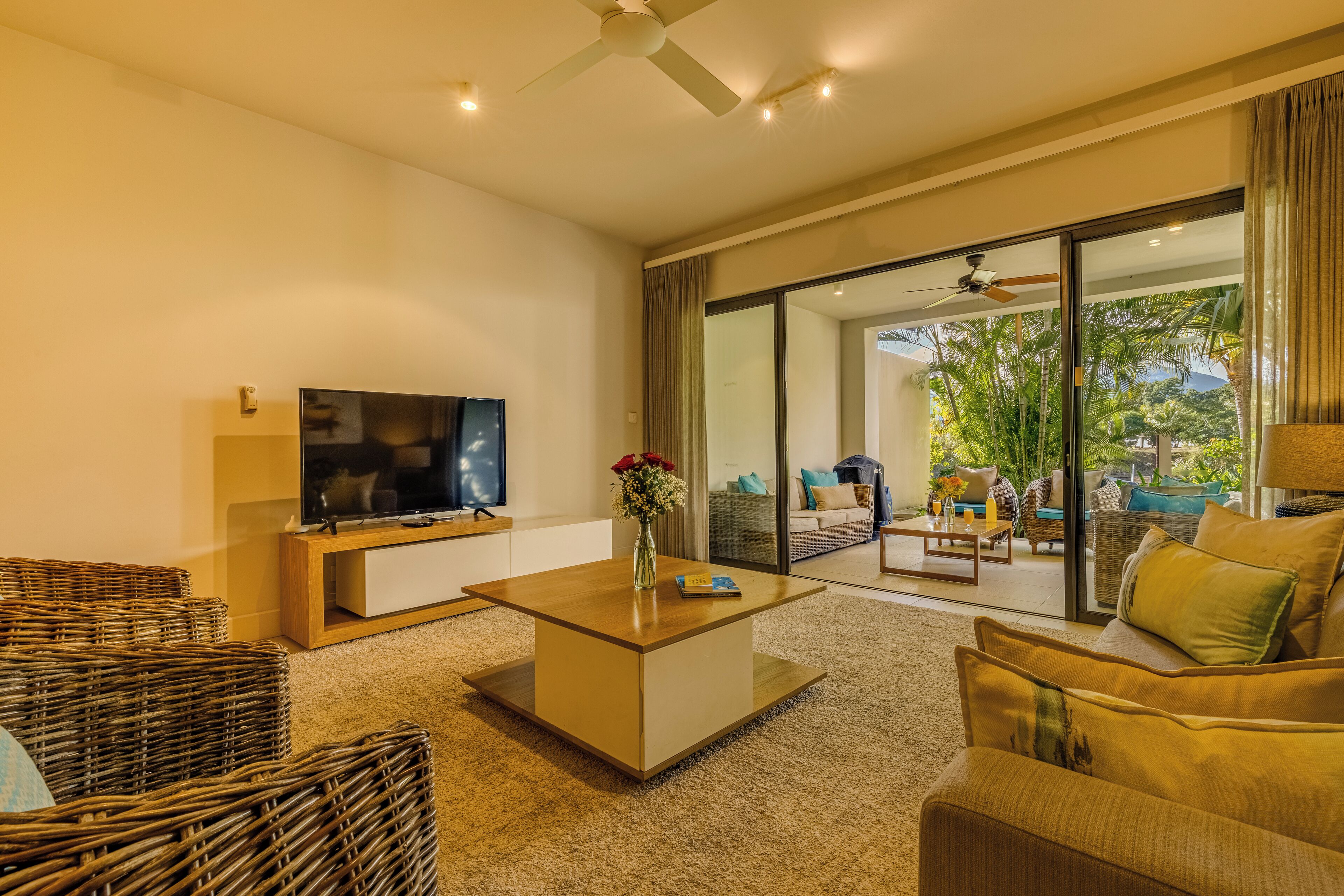 A modern living room with a flat-screen TV, a coffee table, wicker furniture, and a view of the outdoors through large glass doors.