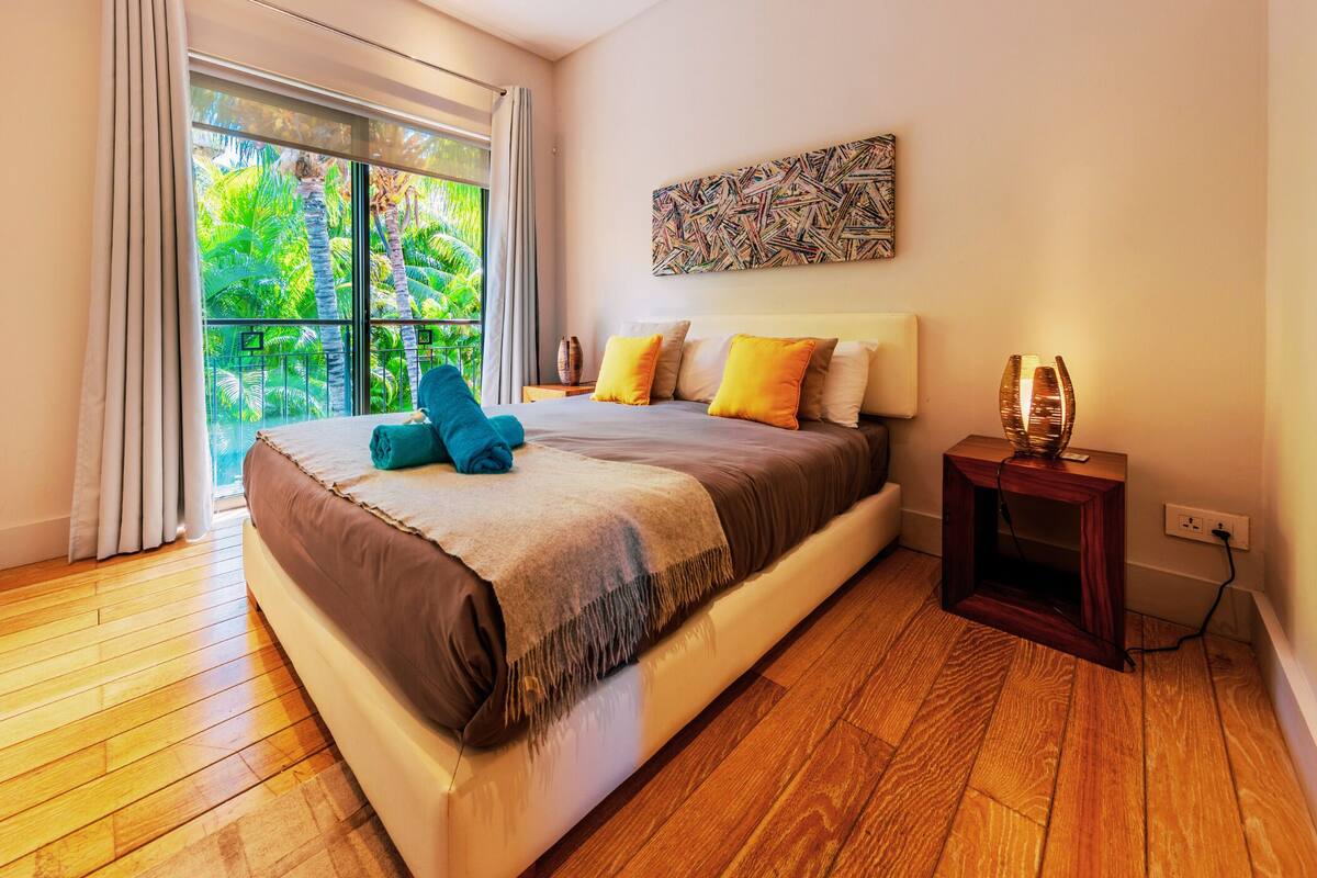 Ein Schlafzimmer mit einem großen Bett, einem Nachttisch, einer Lampe und Blick ins Grüne durch ein Fenster.