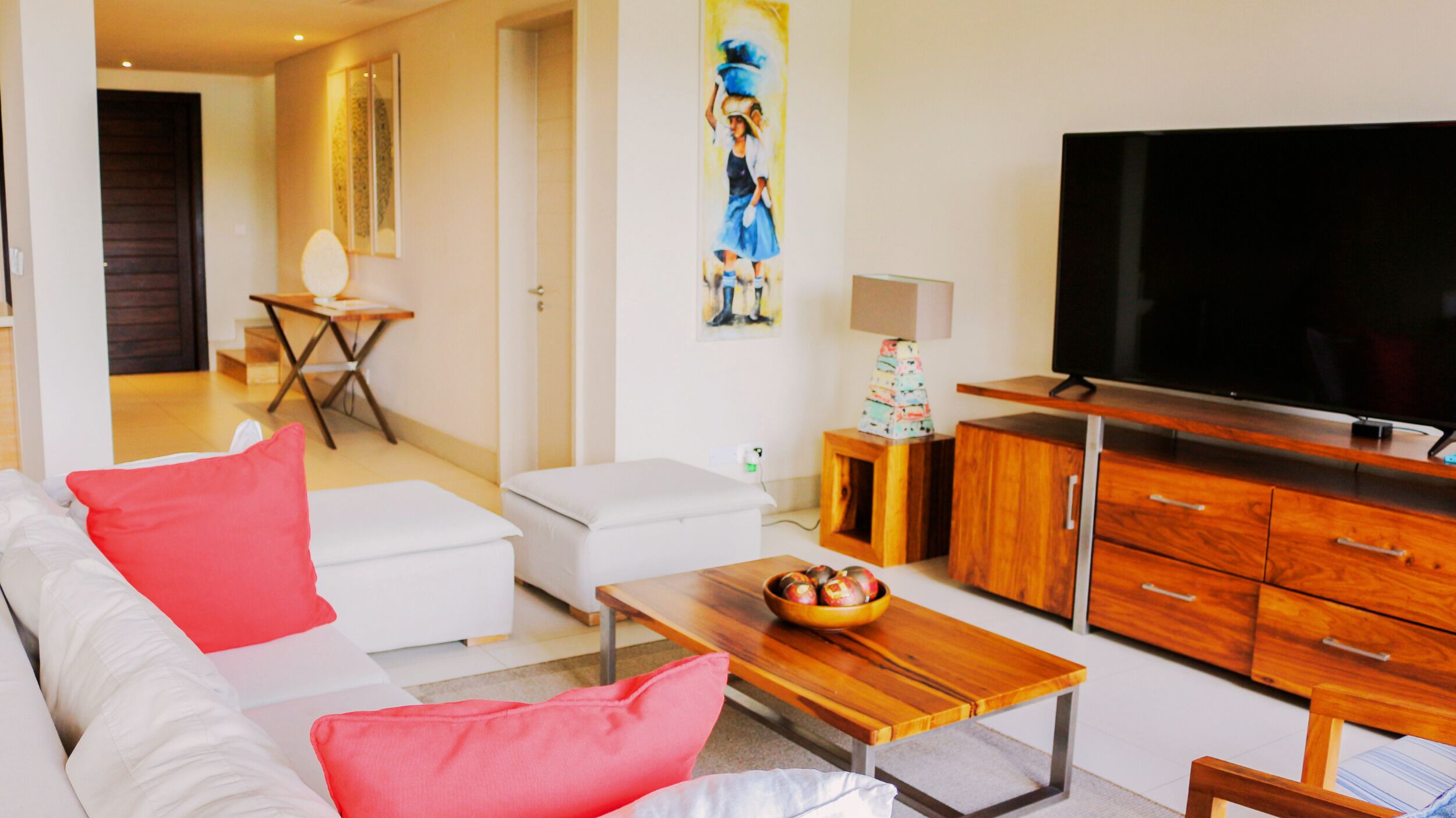 A modern living room with a sofa, ottoman, coffee table, and a TV. There is a wooden cabinet and a small table with a bowl of fruit.