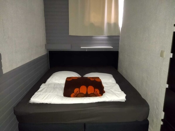 Une petite chambre avec un lit simple, dotée d’une tête de lit sombre, de draps blancs et d’un oreiller marron orné de motifs orange.