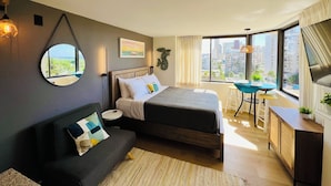 A modern bedroom with a bed, a sofa, a round mirror, and a view of the city through large windows.
