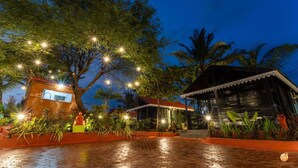 A wooden building with a red roof and a large tree in front, surrounded by plants and lights. - Timber Tide Beach Cottages By Green Concept (Candolim)