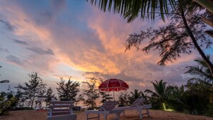 A beach setting with two white lounge chairs and a red umbrella, set against a backdrop of a sunset sky. - Timber Tide Beach Cottages By Green Concept (Candolim)