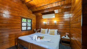 A wooden cabin room with a bed, a window, an air conditioning unit, and a nightstand with a vase. - Timber Tide Beach Cottages By Green Concept (Candolim)