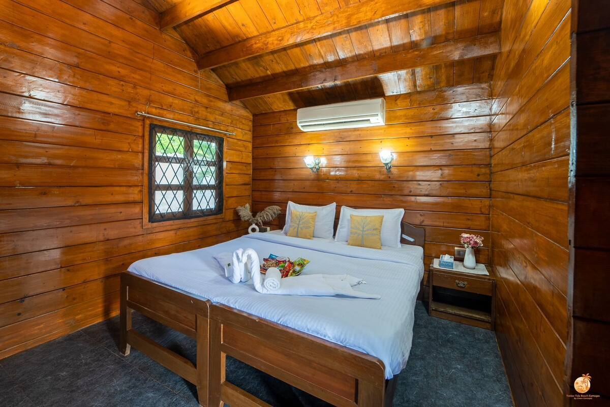 A wooden cabin room with a bed, a window, an air conditioning unit, and a nightstand with a vase.