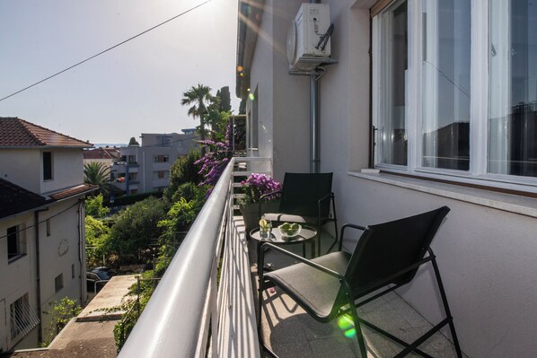 Un balcon avec une table et des chaises, donnant sur un jardin où poussent des fleurs violettes et offrant une vue sur des bâtiments.
