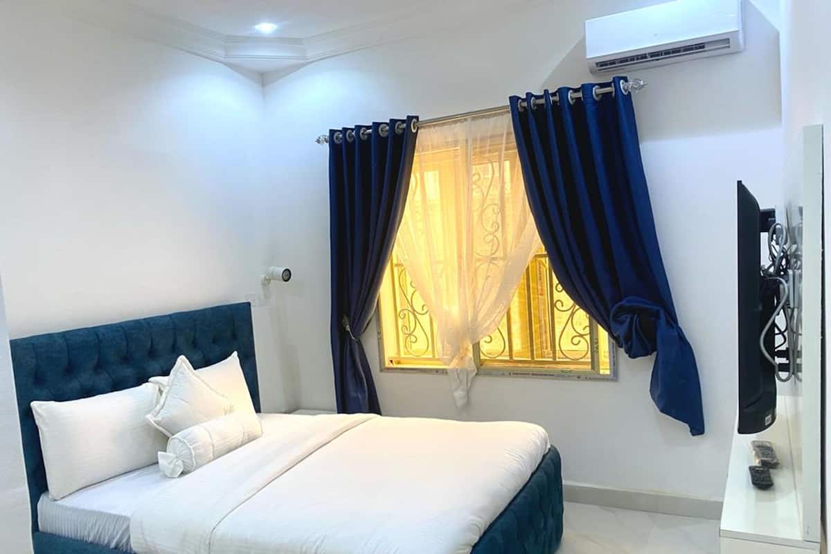 A bedroom with a bed, curtains, a window, an air conditioning unit, and a TV.