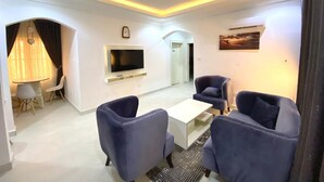 A modern living room with a flat-screen TV, two blue armchairs, a coffee table, and a dining area with a lamp and chairs. - Am Place Luxury Apartments (Abuja)