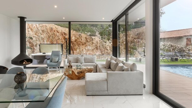 A modern living room with a glass coffee table, a sofa, and a chair. There is a fireplace and a view of a rocky landscape and a pool outside. - Luxe & Charme Résidence (Porto de Mos)