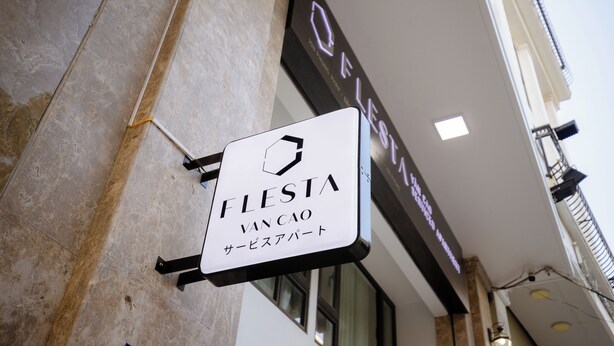 Ein Schild mit der Aufschrift "FLESTA VAN CAO", versehen mit einem sechseckigen Logo, an einem Gebäude mit Steinverkleidung angebracht, oberhalb ein dunkles Schild.