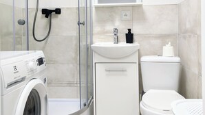 A modern bathroom with a glass-enclosed shower, a washing machine, a sink, and a toilet. - Capsule hostel Floor -1 Self Check in (Warsaw)