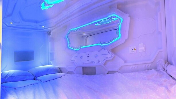 A room with a bed, a wall-mounted control panel, and a power outlet. - Capsule hostel Floor -1 Self Check in (Warsaw)