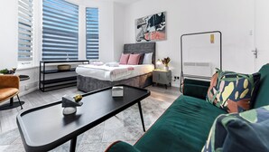 A modern bedroom with a bed, bedside table, and a window with blinds. - Modern Oasis: 2BR Escape with Outdoor Retreat (London)