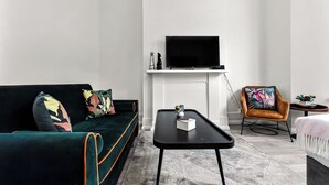 A modern living room with a dark green sofa, a black coffee table, a flat-screen TV mounted above a white fireplace, and a brown armchair with a colorful cushion. - Modern Oasis: 2BR Escape with Outdoor Retreat (London)
