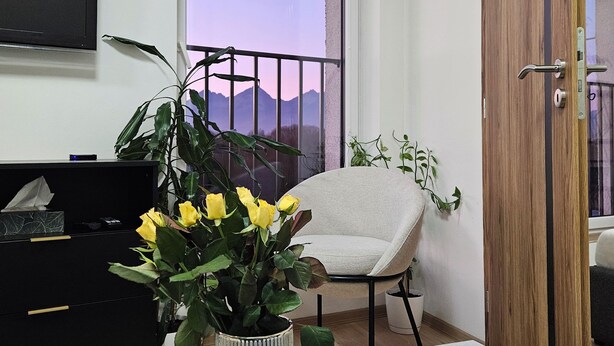 Un soggiorno con una grande finestra, un mobile TV nero, una poltrona beige, una porta in legno e un tavolino in marmo con una candela e una pianta in vaso.