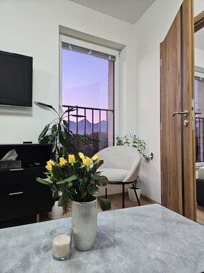 Una sala de estar con un ventanal grande, un mueble negro para entretenimiento, un sillón color beige, una puerta de madera y una mesa de mármol con una vela y una planta en maceta.