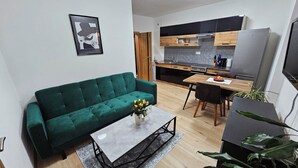 Un ambiente compatto con divano verde, tavolo da pranzo con sedie, angolo cottura con elettrodomestici moderni e una grande pianta in vaso.