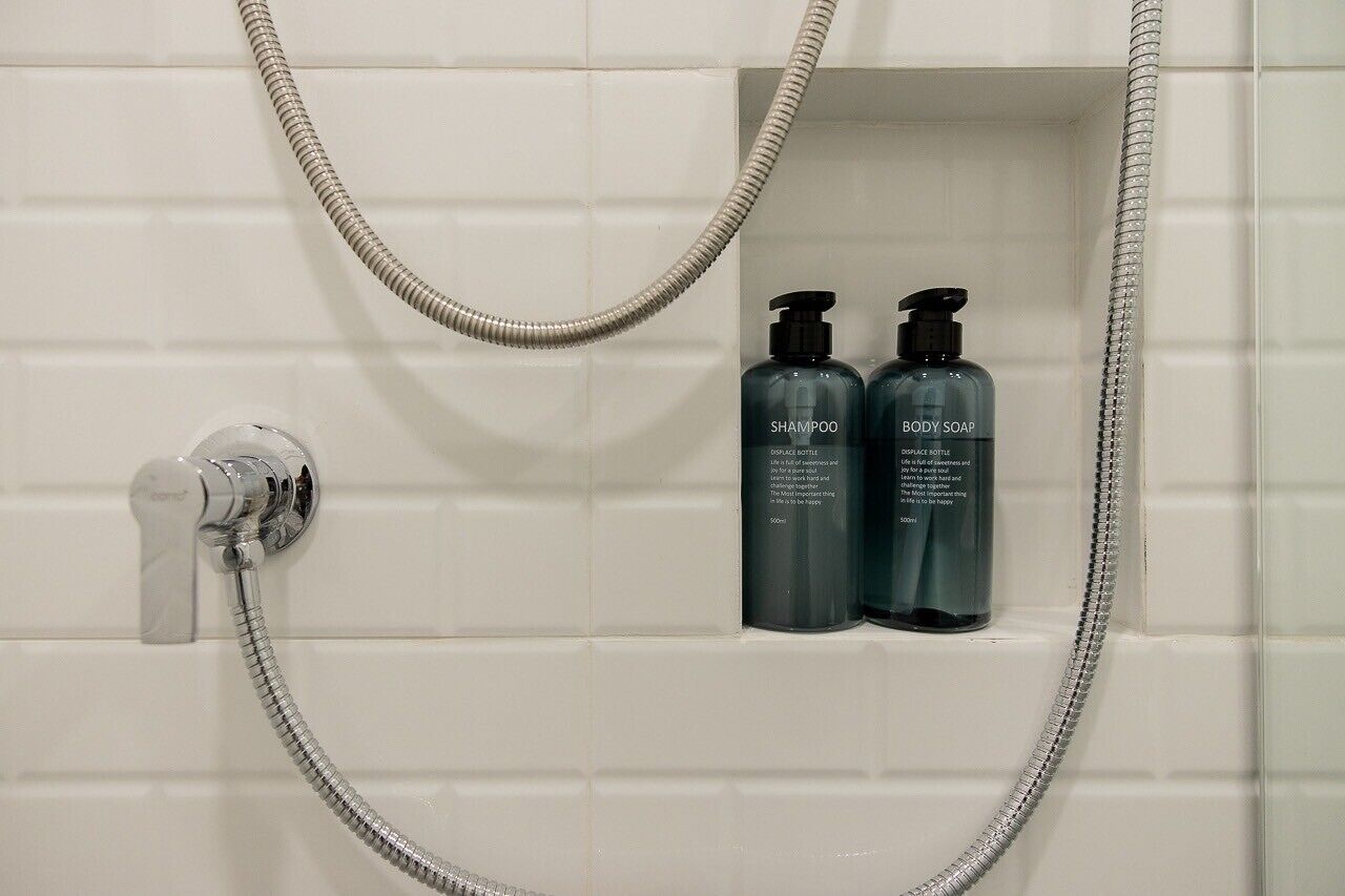 A shower area with a chrome showerhead and two bottles of body soap labeled "SHAMPOO" and "BODY SOAP" in a white tiled enclosure.