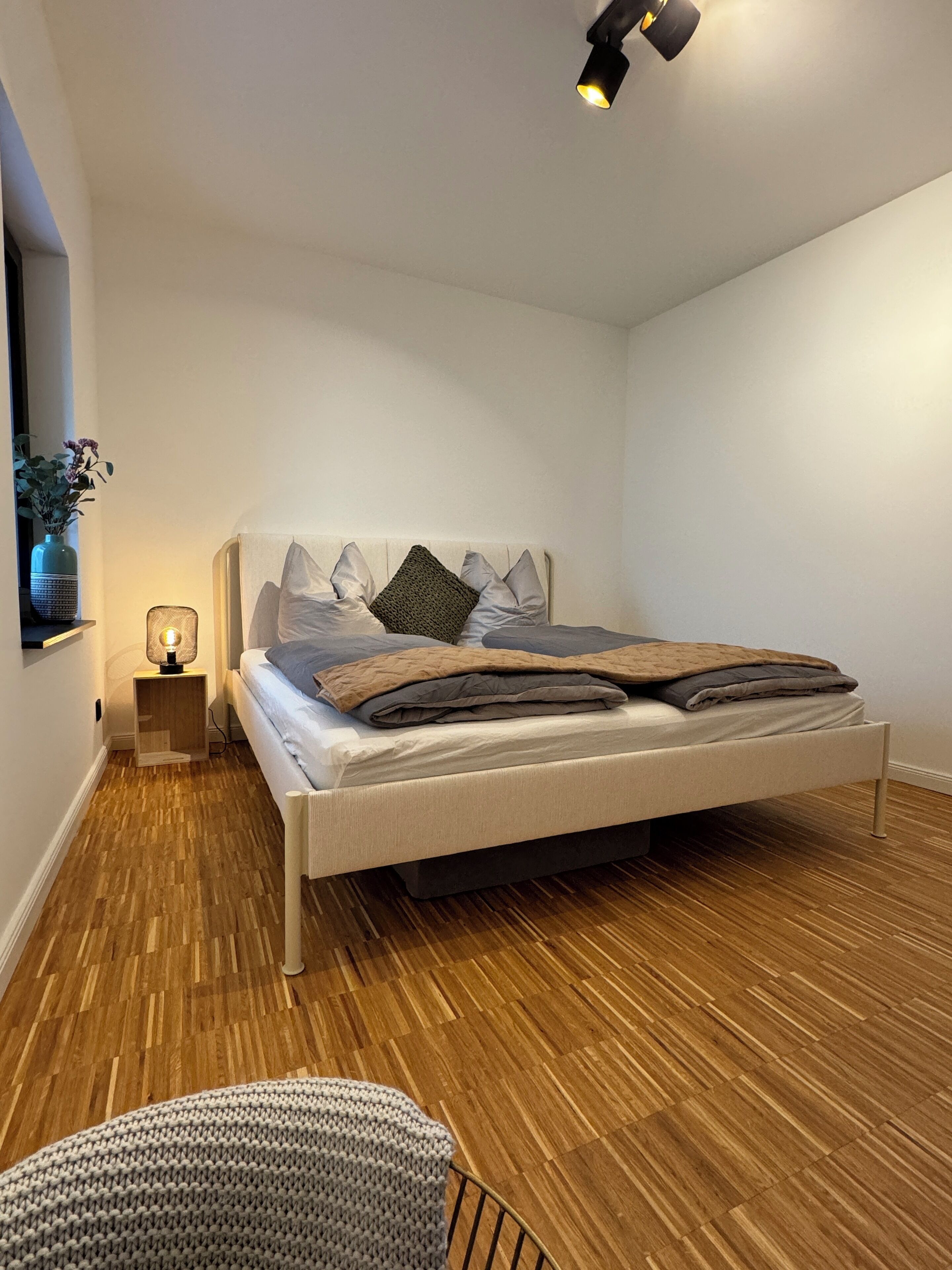 Ein modernes Schlafzimmer mit Holzboden, einem Bett mit grauer und brauner Decke, einer Nachttischlampe und einem kleinen Tisch mit einer Blumenvase.