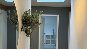 Un couloir avec un grand miroir, une plante en pot et une vue sur une pièce où l’on aperçoit un meuble blanc et un évier.