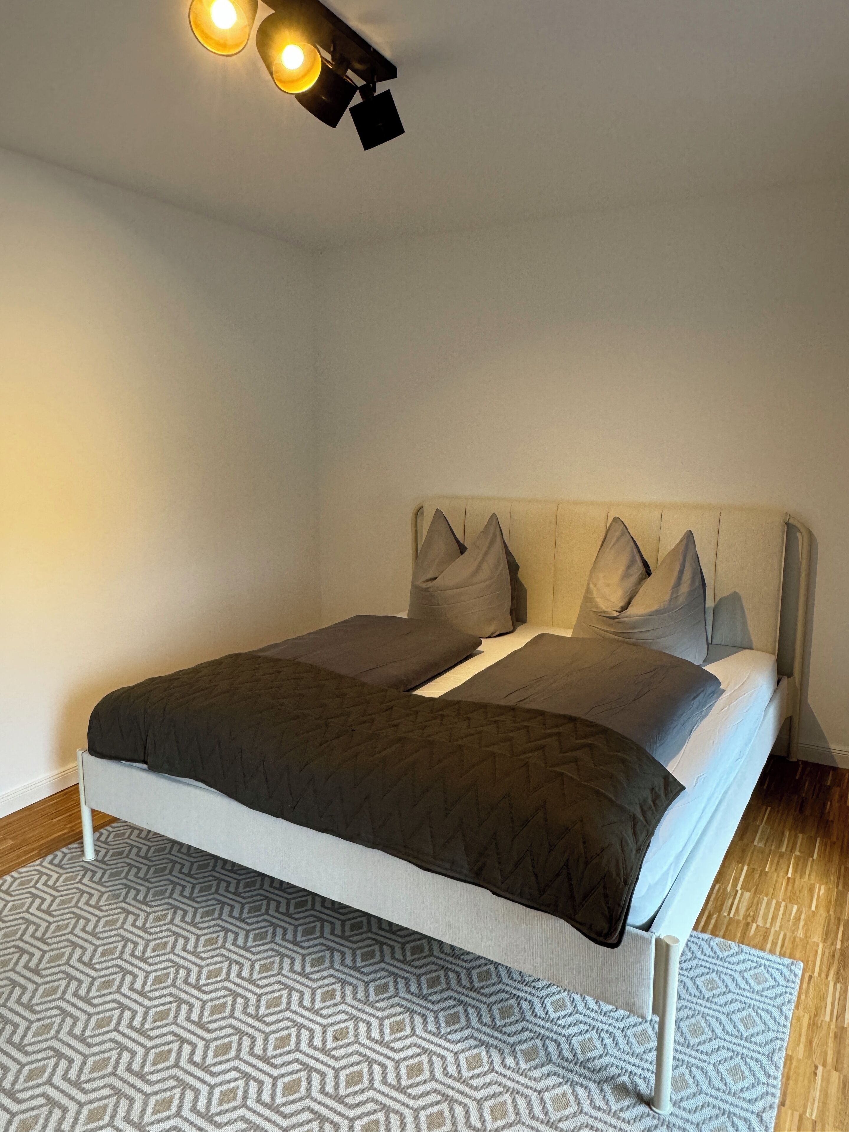 A neatly made bed with a grey and white patterned rug, a light-colored headboard, and a ceiling light fixture.