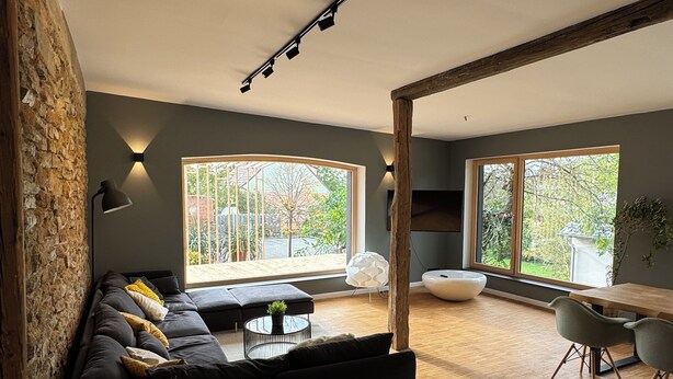 Un salon moderne avec un canapé d’angle noir, une table basse ronde et un coin repas comprenant une table et des chaises en bois. De grandes fenêtres laissent entrer la lumière naturelle et offrent une vue sur la verdure environnante.