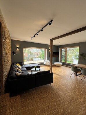 A modern living room with a black sectional sofa, a round coffee table, and a dining area with a wooden table and chairs. Large windows provide natural light and views of greenery outside.