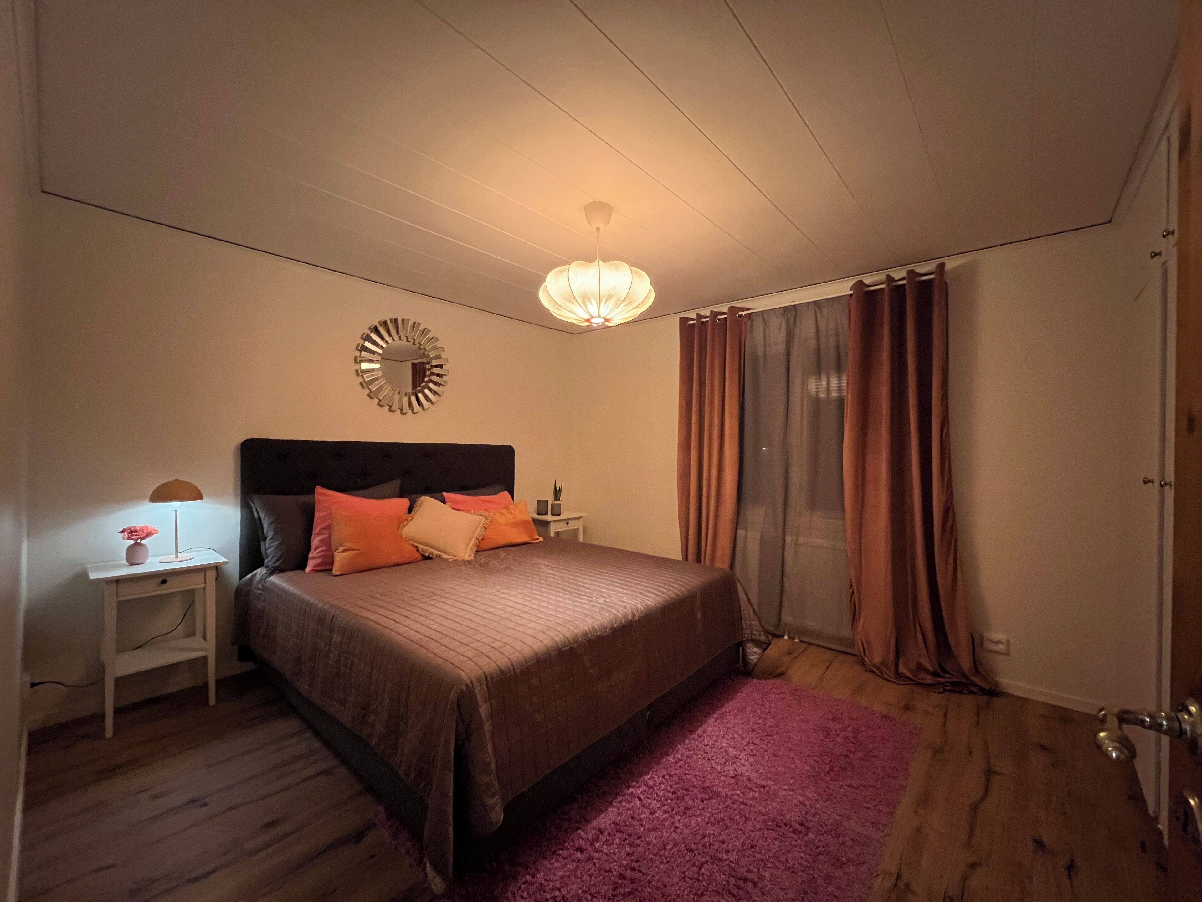 A bedroom with a bed, nightstand, lamp, mirror, and curtains.