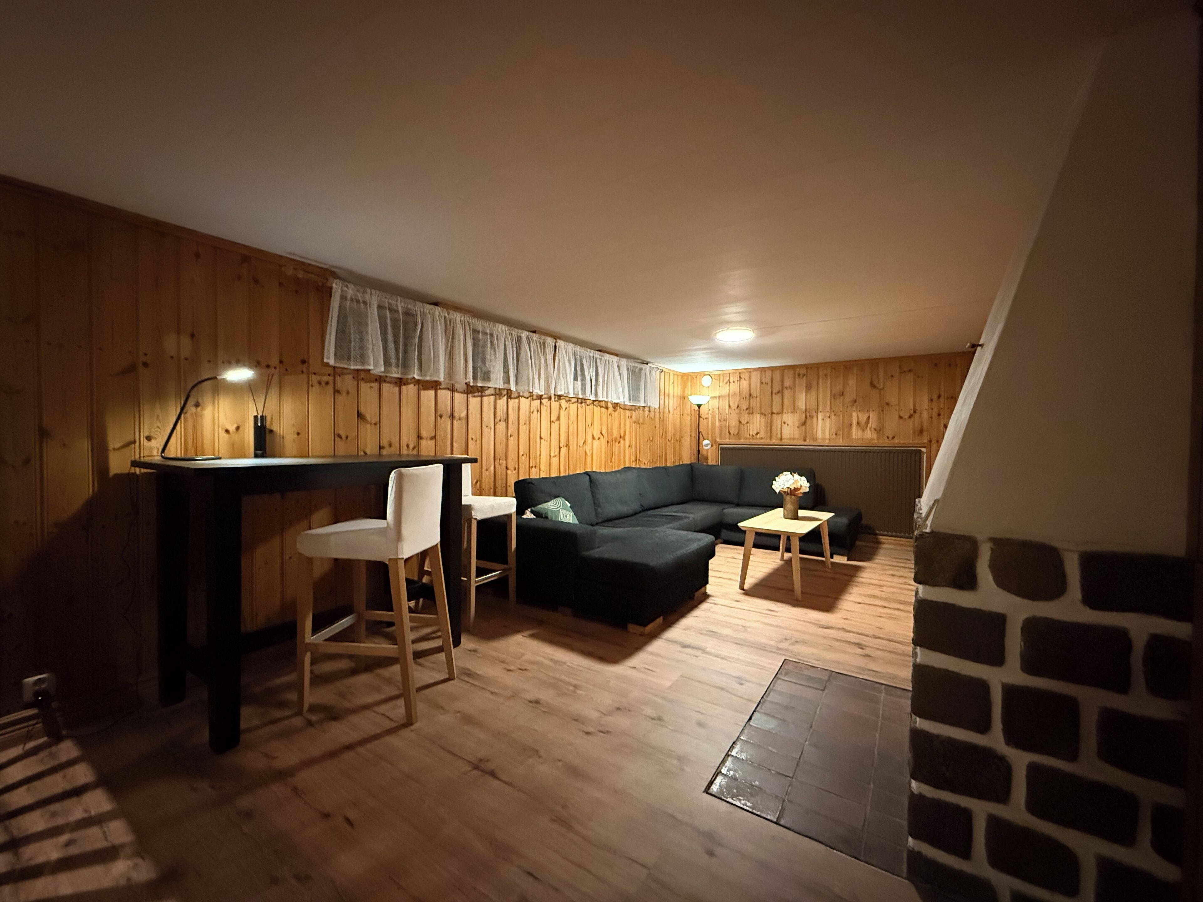 Una habitación acogedora con piso de madera, un sofá negro, una pequeña mesa con un florero y un escritorio con dos sillas.