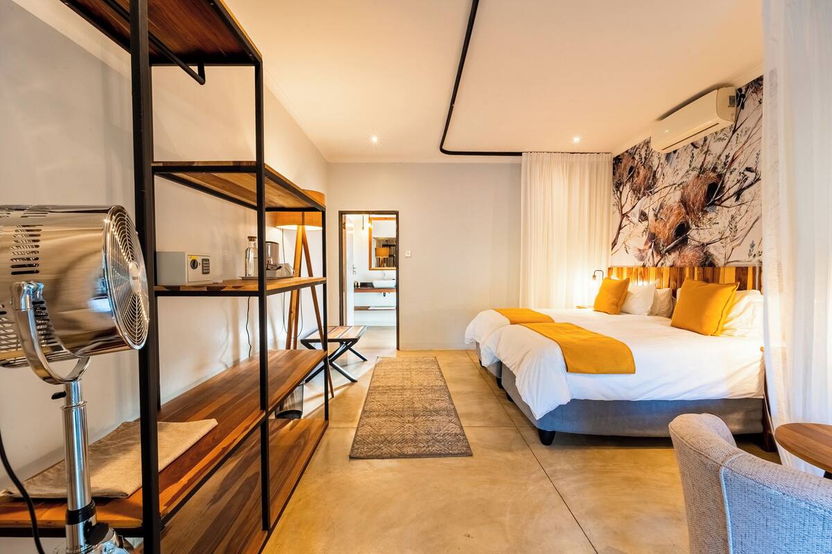 Quarto de hotel moderno com uma cama grande, um banco de madeira, um ventilador e vista para o corredor.