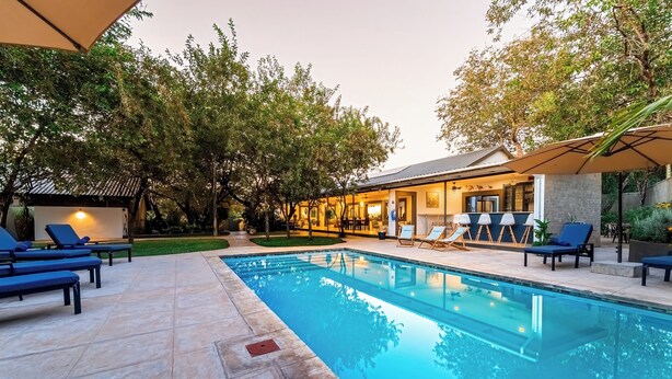 A swimming pool with lounge chairs and umbrellas, surrounded by trees and a building with outdoor seating. - Kabakwe Guest Lodge (Victoria Falls)