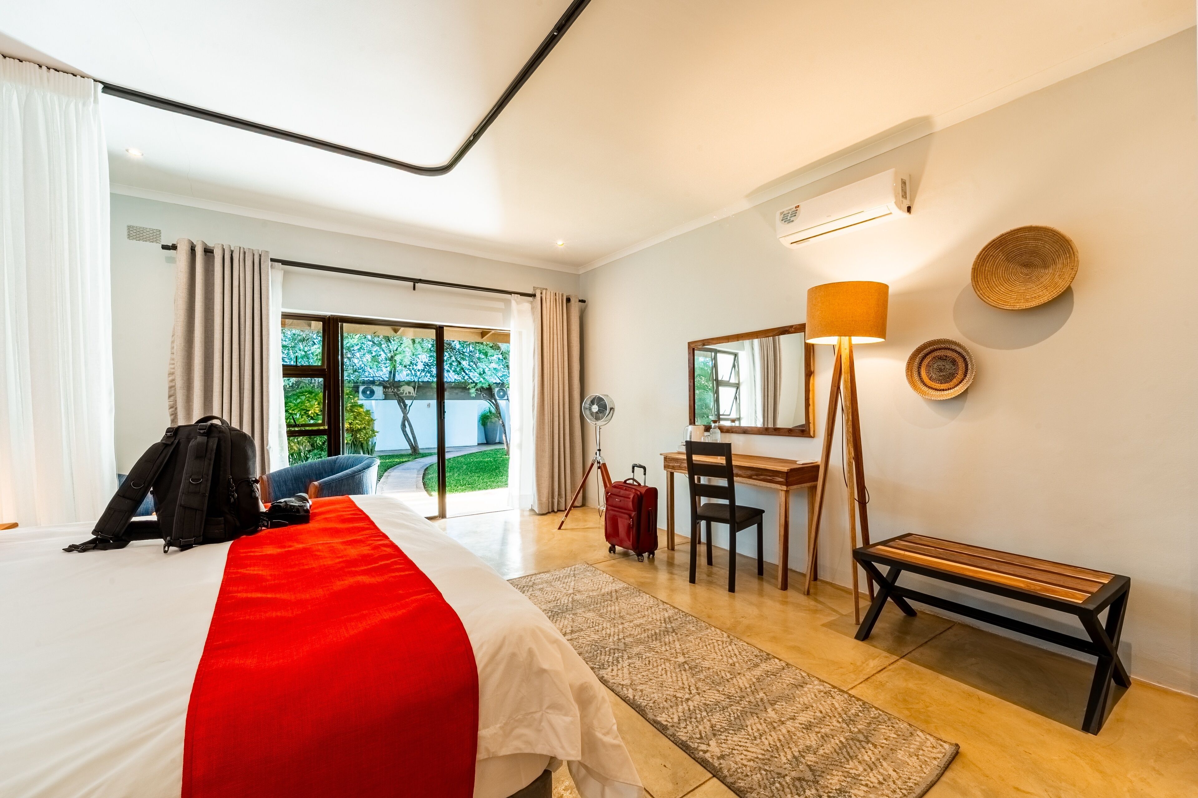 A hotel room with a bed, a desk, a chair, a bench, a lamp, and a view of the outdoors.