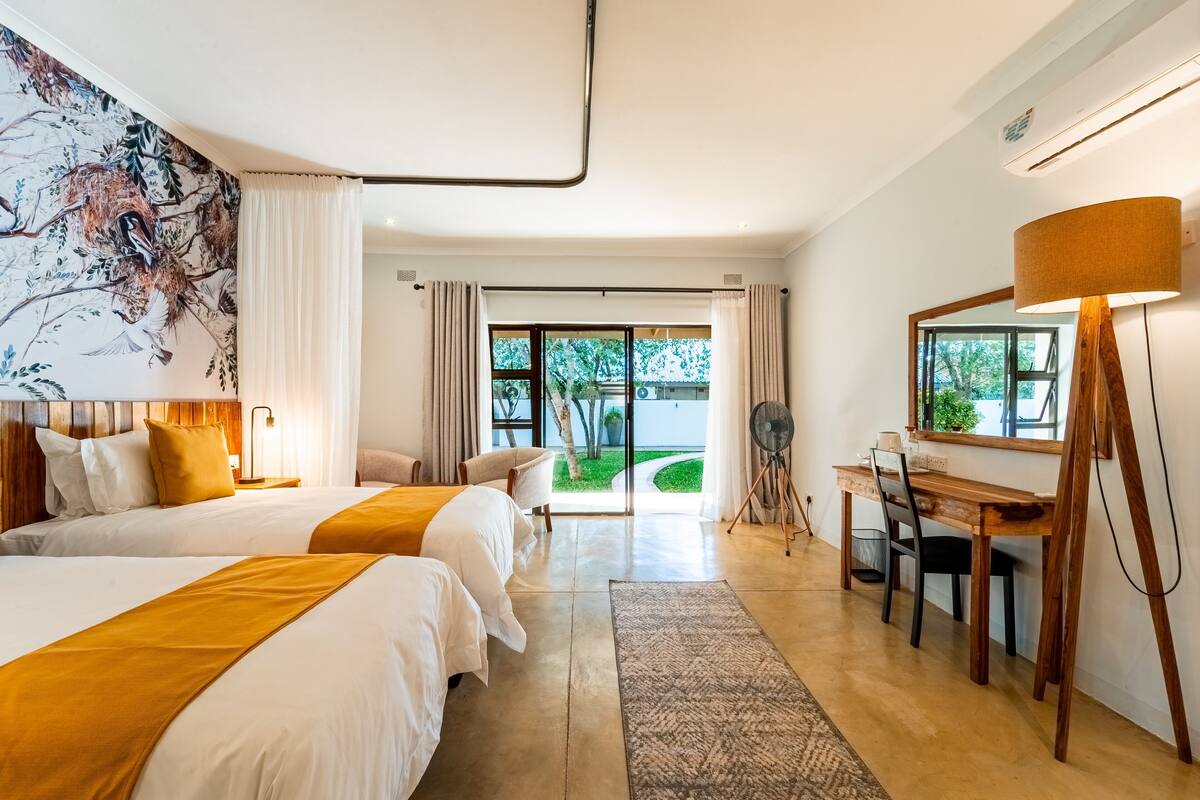 Quarto de hotel com duas camas, uma escrivaninha, uma cadeira, uma luminária e uma janela grande com vista para vegetação.