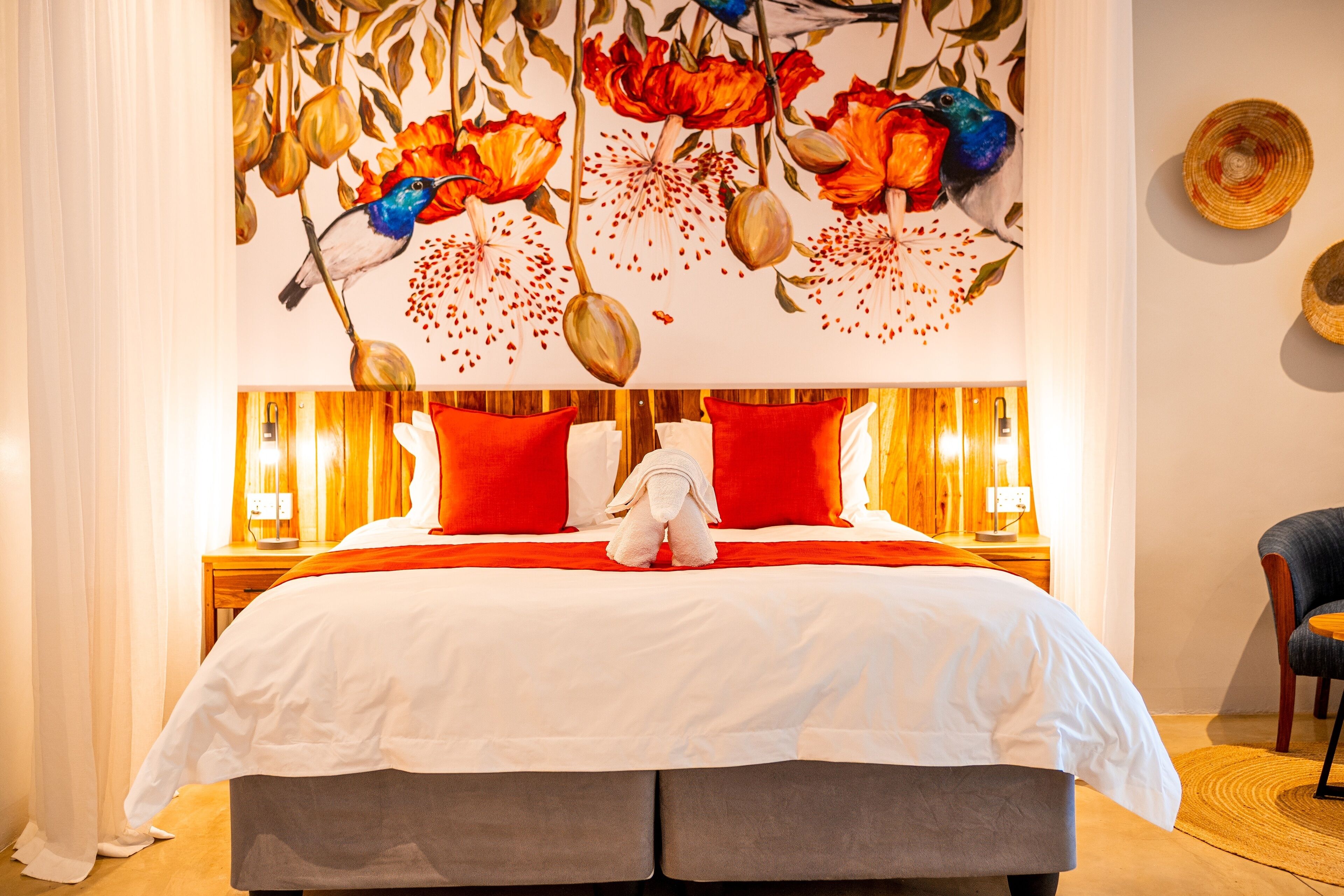 A hotel room with a large bed, red and white bedding, a decorative wall mural, and a wooden nightstand with a lamp.