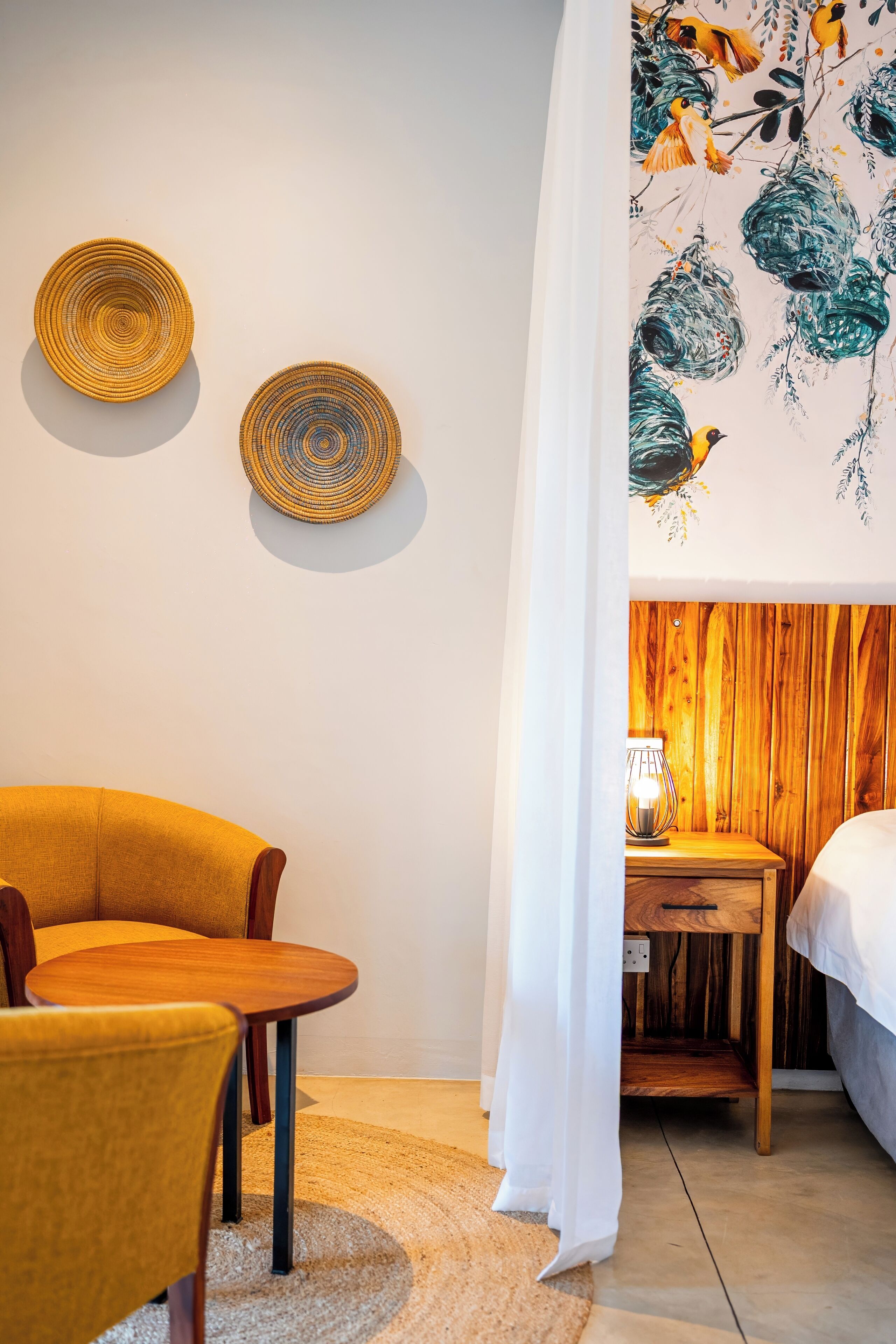 A room with a yellow armchair, a round wooden table, a wall-mounted woven basket, and a bed with a wooden headboard.