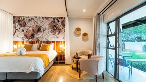 A modern hotel room with a large bed, a seating area with a table, and a view of the outdoors through large windows. - Kabakwe Guest Lodge (Victoria Falls)