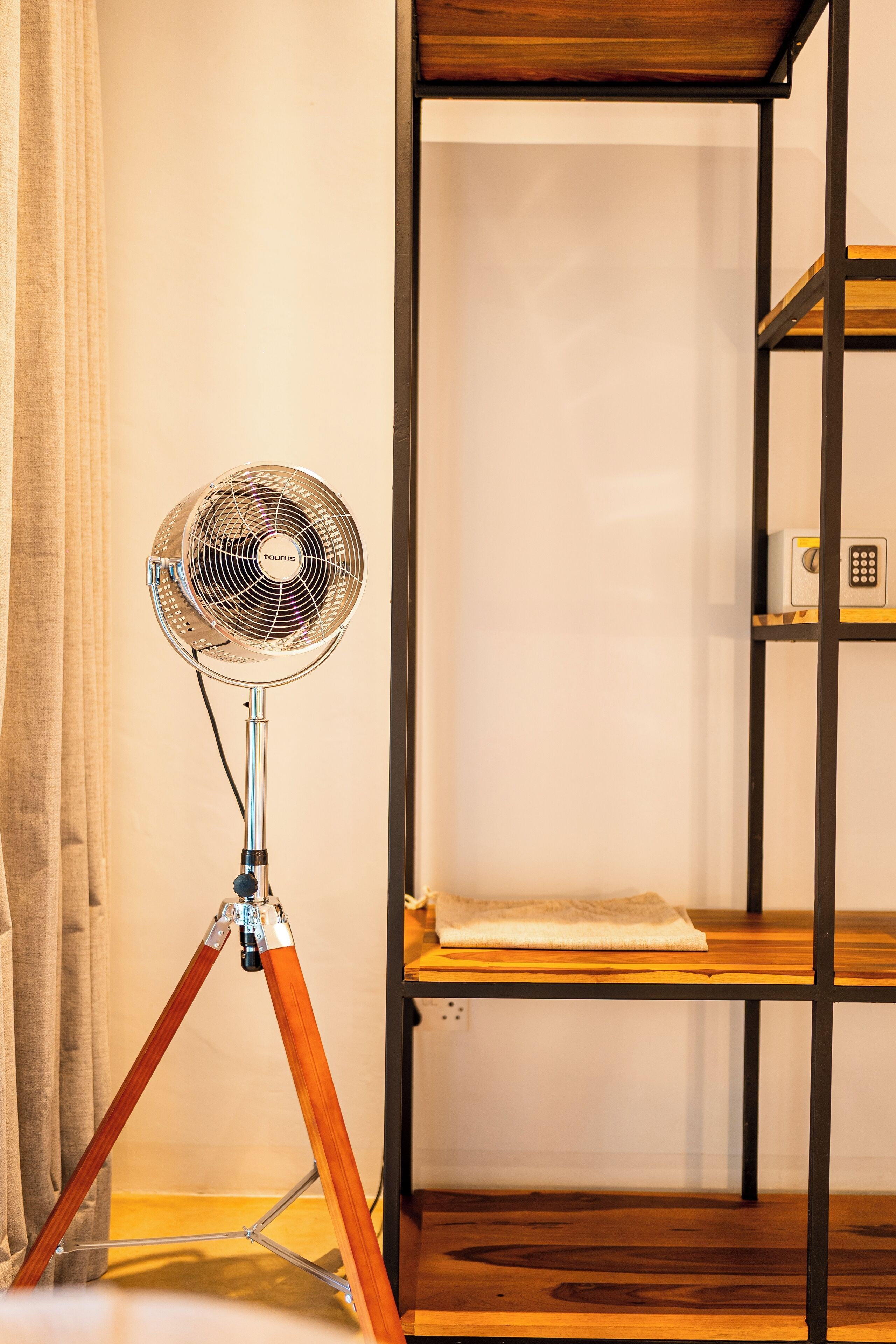 A room with a standing fan on a tripod, a wooden shelf unit, and a curtain.