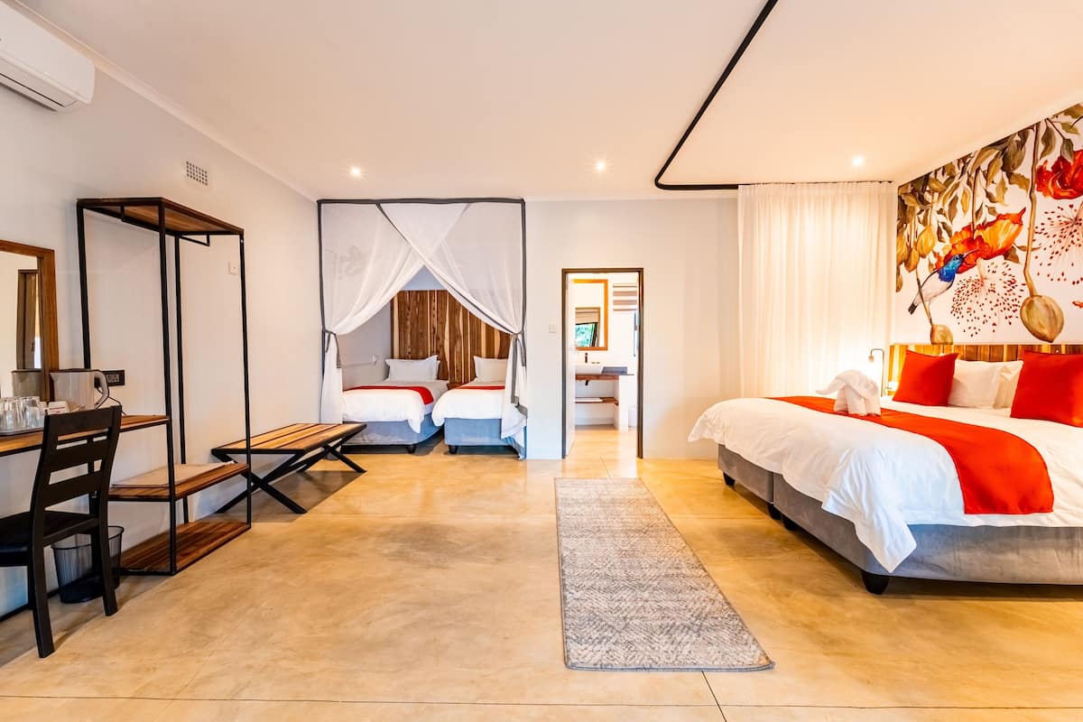 Quarto de hotel moderno com uma cama grande, uma escrivaninha com cadeira, uma mesinha e um banco. Um banheiro é visível através de uma porta aberta.