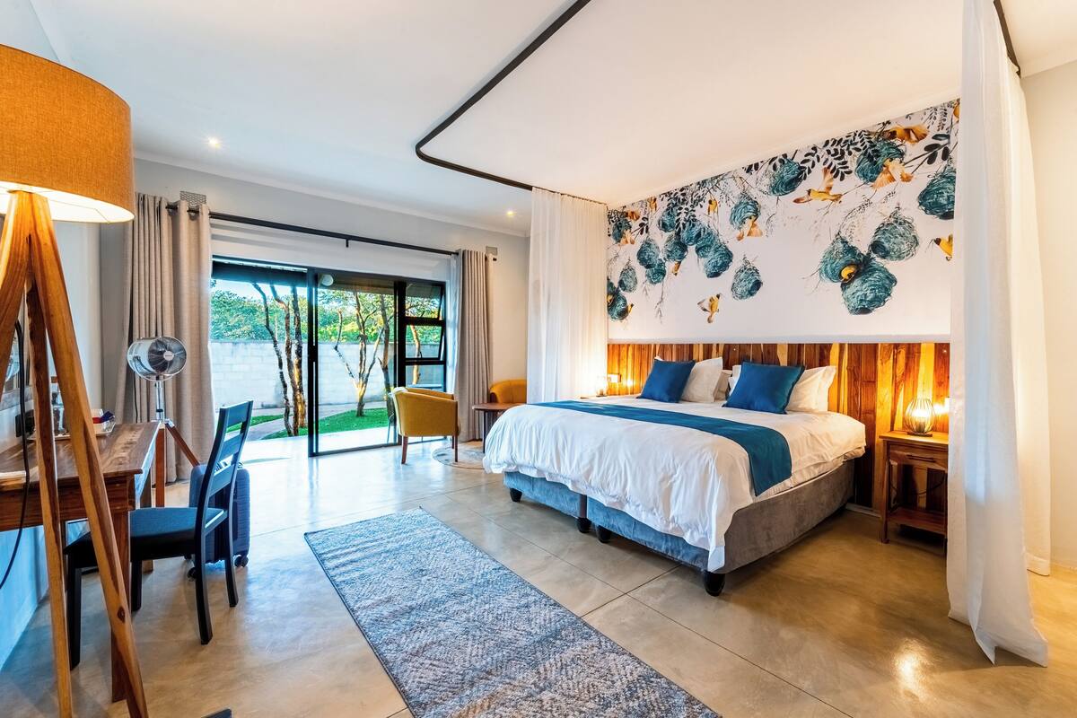 Quarto de hotel moderno com uma cama grande, uma escrivaninha com cadeira, um abajur e vista para o exterior.
