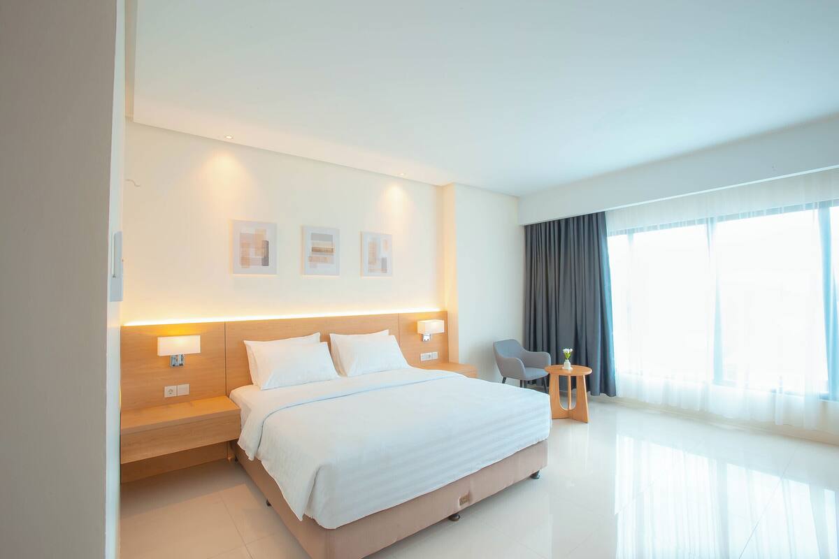 Quarto de hotel moderno com uma cama grande, abajures, uma mesinha e uma cadeira.