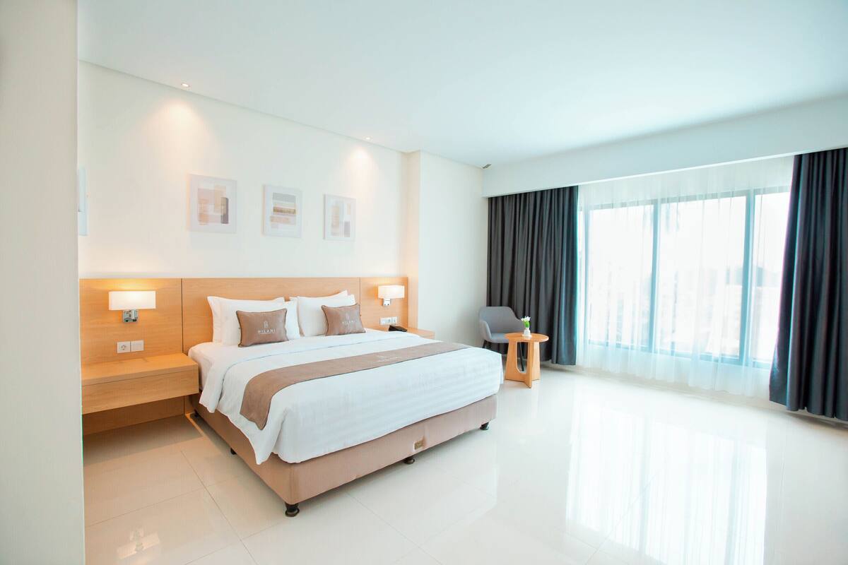 Quarto de hotel moderno com uma cama grande, abajures, uma mesinha e vista para o exterior através de amplas janelas.