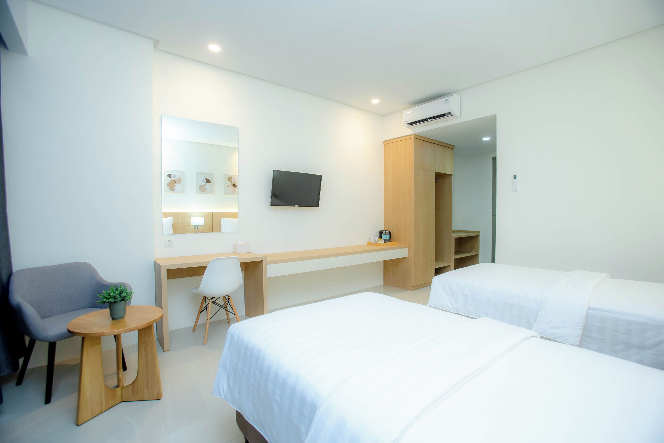 A modern hotel room with two beds, a desk, a chair, a small table with a plant, a TV mounted on the wall, and an air conditioning unit.