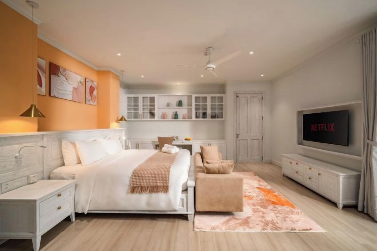 Un dormitorio moderno con cama, mesitas de noche, un sofá, un televisor y una cocina al fondo.