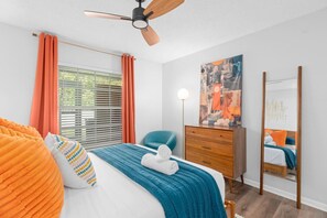 Un dormitorio con cama, cómoda, silla, ventilador de techo, ventana con cortinas y un cuadro en la pared.