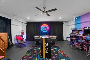 Una habitación con mesa de billar, máquinas de juego, ventilador de techo y letrero de neón.