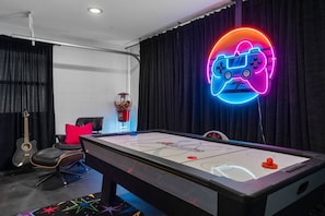 Una mesa de billar con un letrero luminoso de control de videojuegos, una guitarra y una almohada roja en una habitación con cortinas negras y una pared blanca.