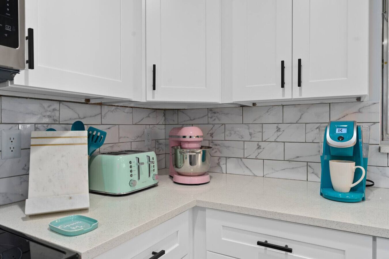 A kitchen with white cabinets, a turquoise coffee maker, a pink mixer, a teal toaster, and a white mug.